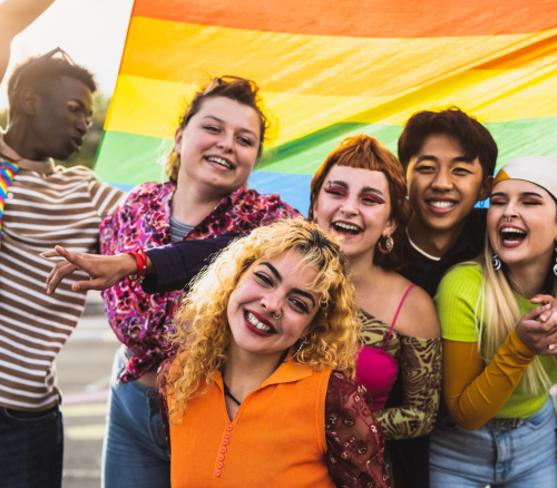 A group of teenagers of mixed genders and ethnicities smiling and laughing while holding the LGBTQ+ flag.